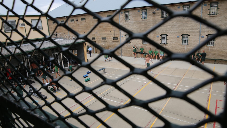 Inmates wearing green clothes exercise in a yard as seen through a fence.