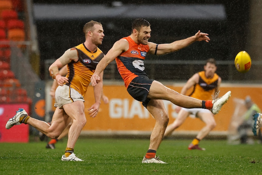 A GWS player extends his foot as he kicks the ball downfield in the rain as a Hawthorn player runs behind him. 