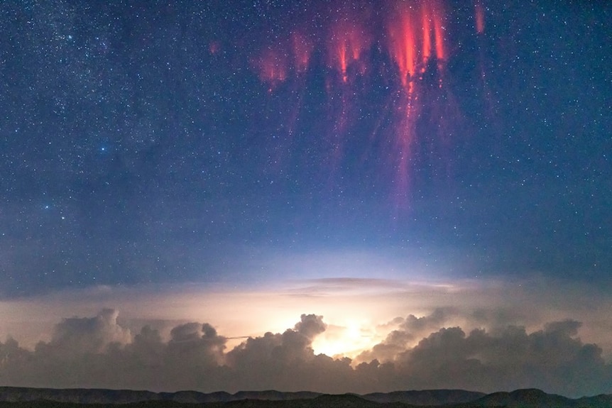 Red streaks are seen in a dark starry sky above storm clouds hovering above a mountainous desert landscape.