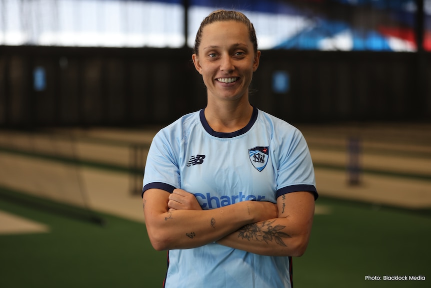 A woman smiles standing with her arms crossed wearing a blue jersey in front of indoor cricket nets.