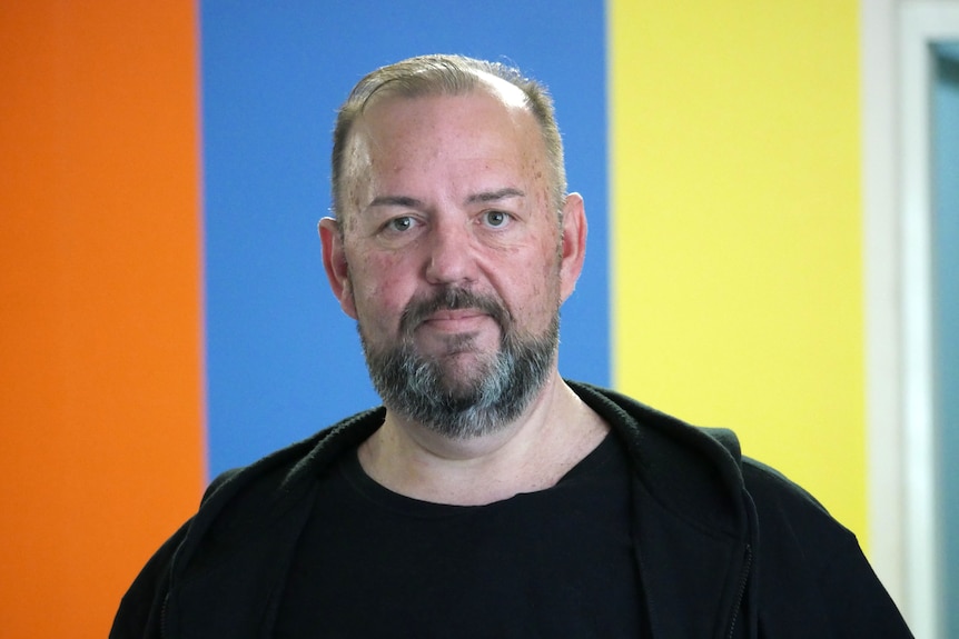 A man looking down the camera with a colourful wall behind him.