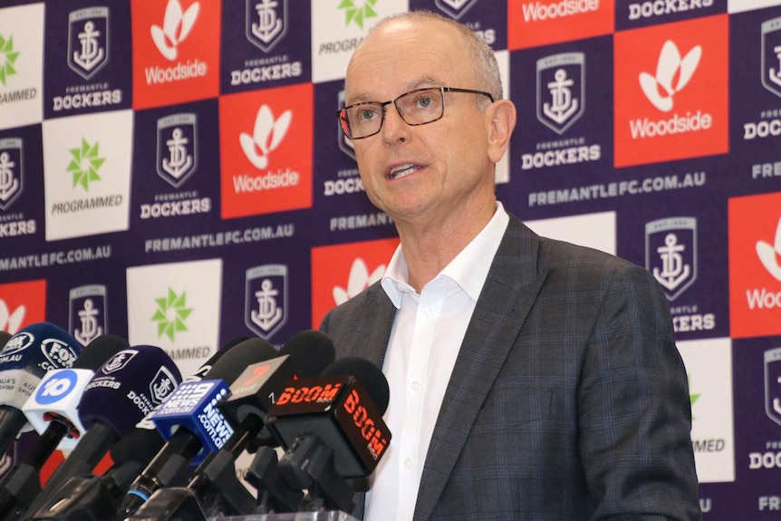 A mid-shot of Dale Alcock speaking in front of a board of Fremantle Dockers sponsor logos.