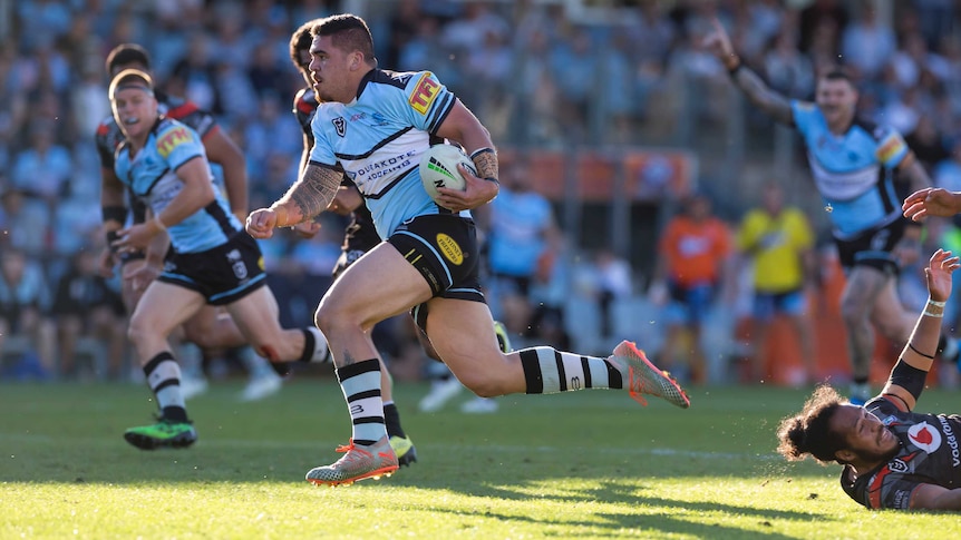 A male NRL player runs the ball in his left hand as he looks to score a try.