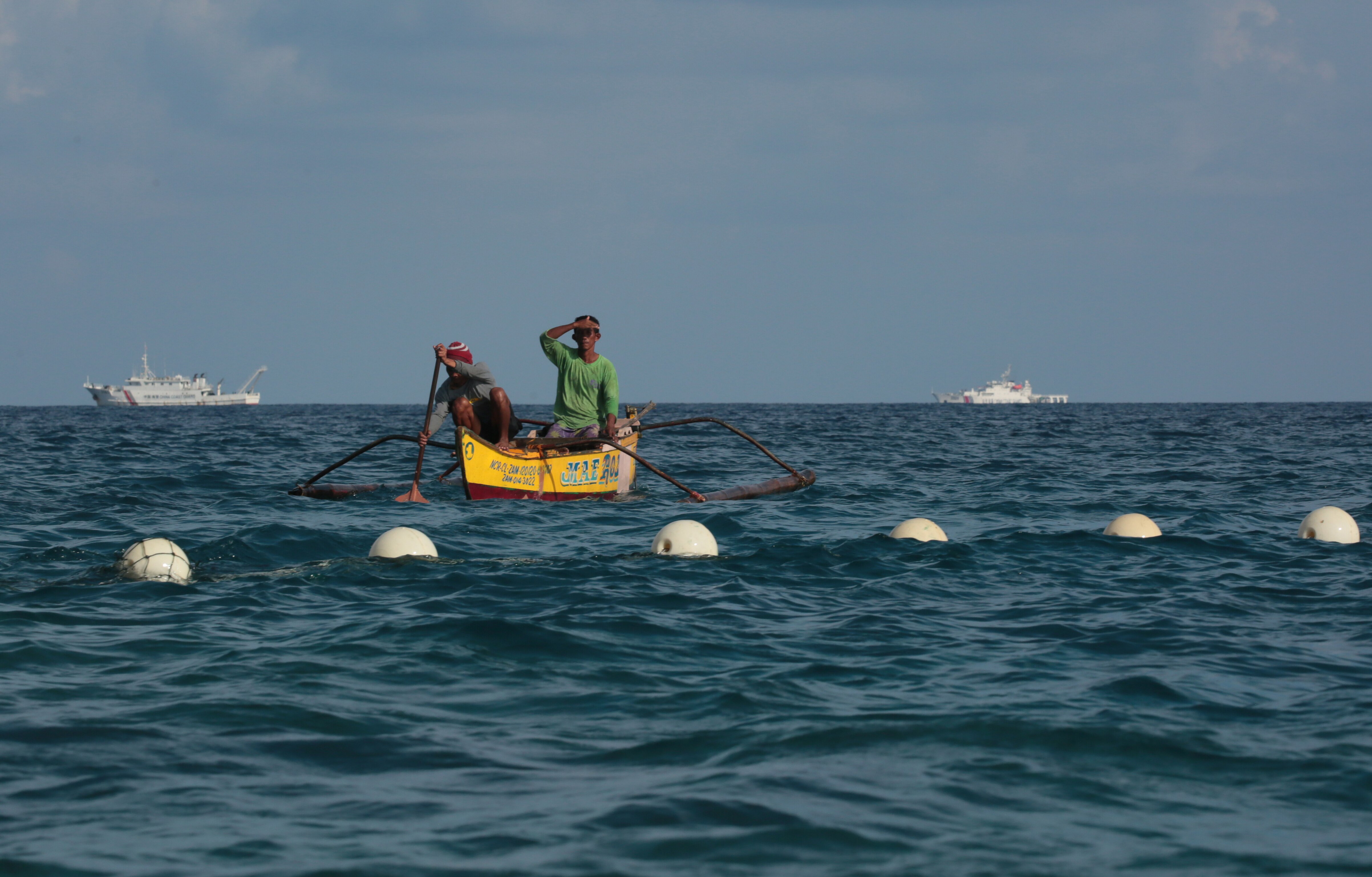 Two people in a boat near a line of buoys with big ships in the distance