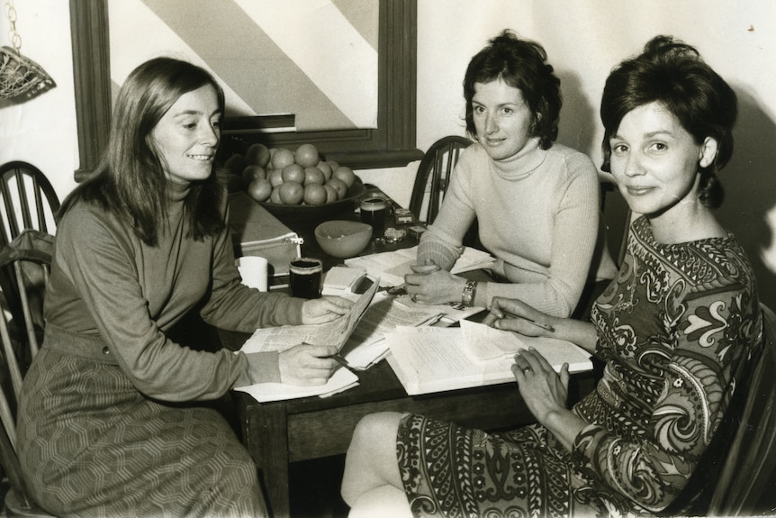 A b&w image of a three women sitting around a dining table looking at papers.