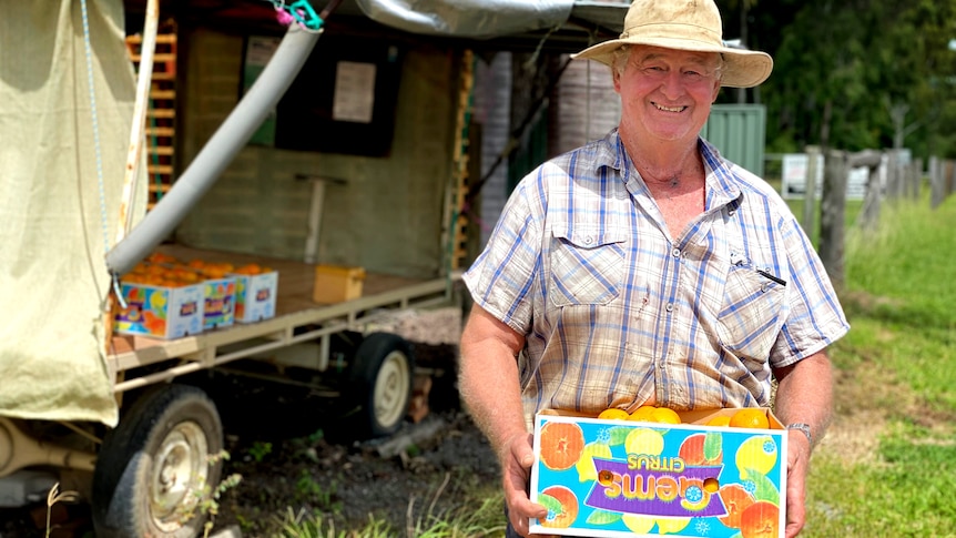 A man wearing farm gear carries a box of mandarins with a roadside fruit stall behind him.