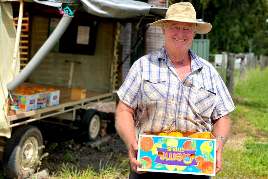 A man wearing farm gear carries a box of mandarins with a roadside fruit stall behind him.