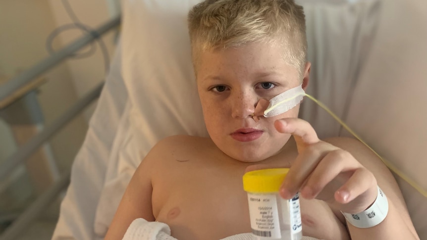 Boy with blonde hair in hospital bed with a tube coming out of his nose, holding a specimen jar with a gaming console