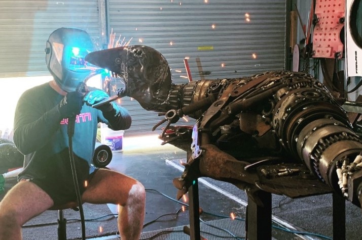 Steve Ross in a long sleeve shirt and footy short welding with with a mask on and sparks flying off a metal sculpture dinosaur.
