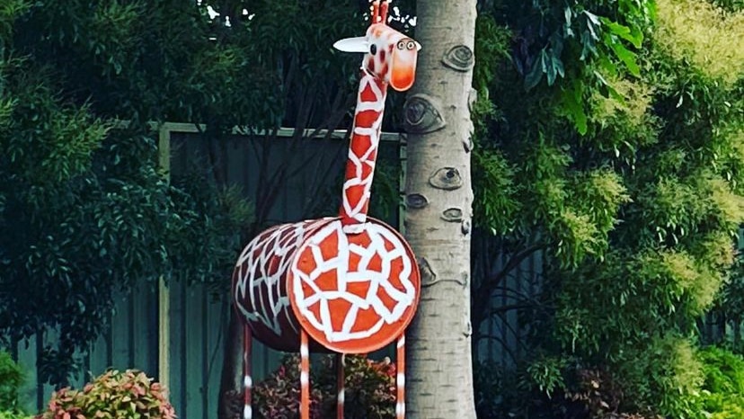 Jerry the giraffe tucked amongst a palm tree and bushes on a verdant nature strip