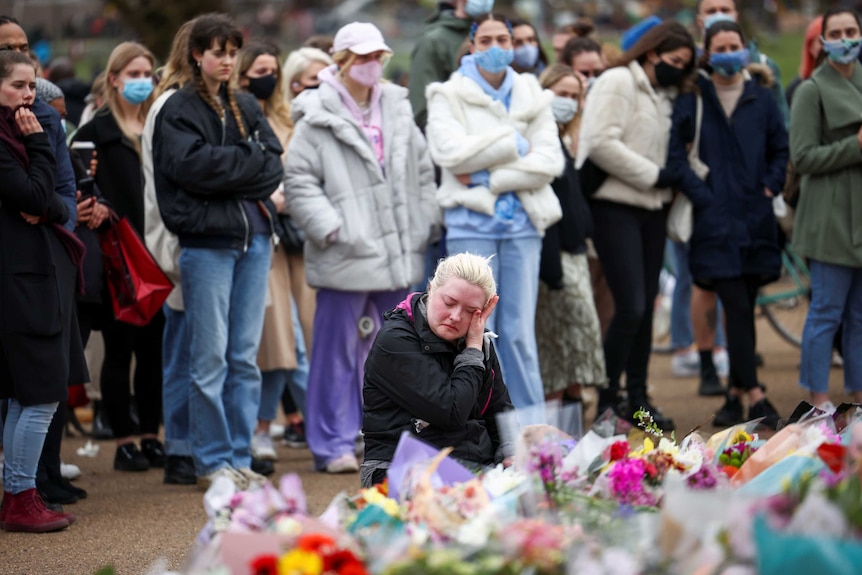 A crouching woman wipes tears from her eye as a crowd of mourners solemnly observe a floral memorial.