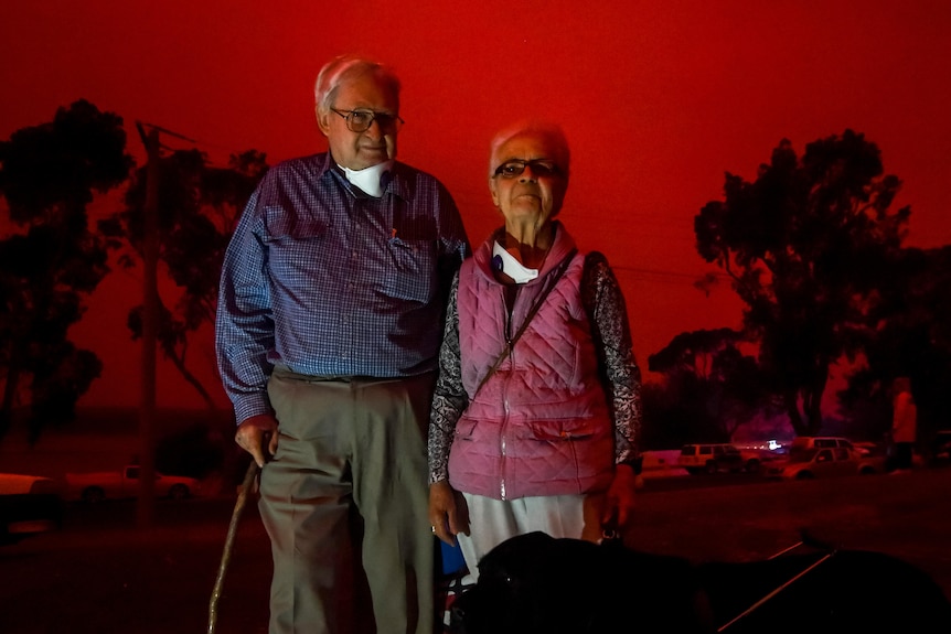 An old couple stands outside with gum trees behind them and the sky completely red.