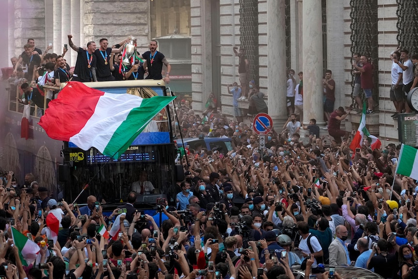 Medal-wearing Italian footballers travel in an open-top bus, swaying and holding Euro 2020 trophy as fans wave Italian flags.