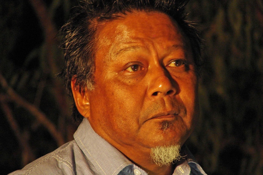 A close up on a man's face against a dark background at night. He wears a serious expression.