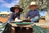 Two boys sit outside with books wearing cowboy hats, jeans and smiling