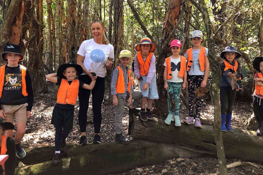Children wearing bright orange vests stand on a log surrounded by trees, smiling and holding hands with an educator.