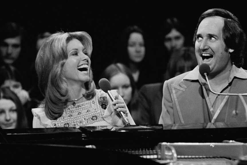 Olivia Newton-John smiles while singing at a piano alongside a crazed-looking Neil Sedaka