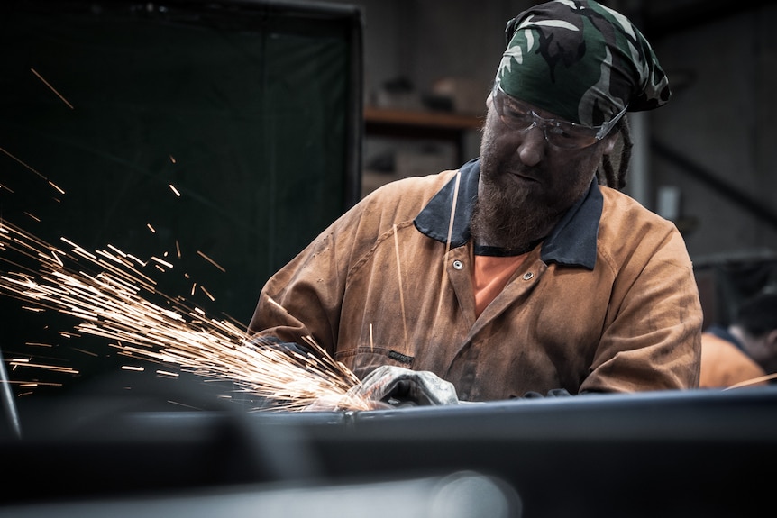 A man with dreadlocks and a bandana uses welding tools, sending sparks flying