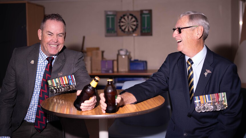 Two men in their army medals sit at a table and cheers with two beers.