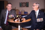 Two men in their army medals sit at a table and cheers with two beers.