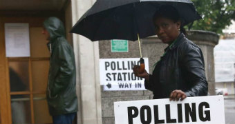 A woman carries a polling station sign and a black umbrella