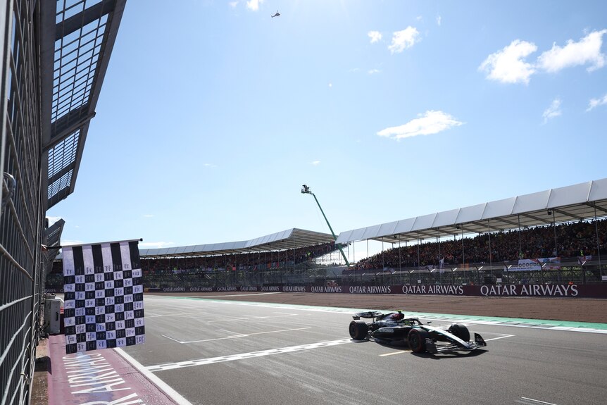 The chequered flag is shown as a F1 racing car passes the finish line with a grandstand in the background.