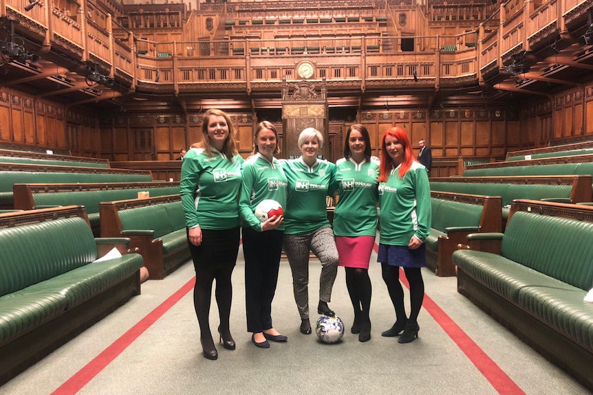 Five UK MPs sport green jerseys as they stand on the floor of the House of Commons, iconic green benches at either side of them.