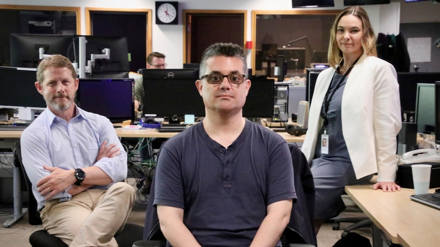 Three journalists sit in a newsroom posing for a photo