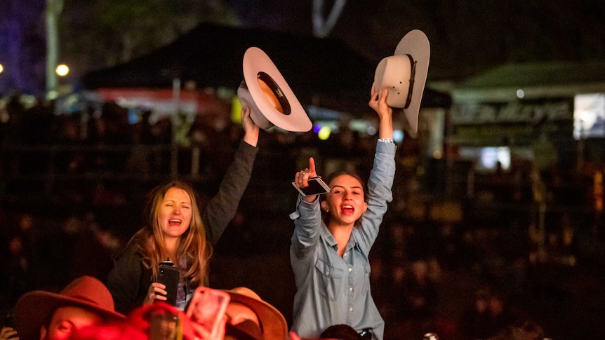 Two girls sitting on someones shoulders rise above a concert crowd waving cowboy hats