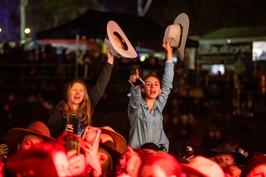 Two girls sitting on someones shoulders rise above a concert crowd waving cowboy hats