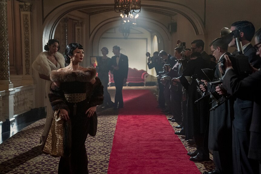 A woman in fur lined evening gown stands and poses for press photographers on red carpet in ornate hazy interior.