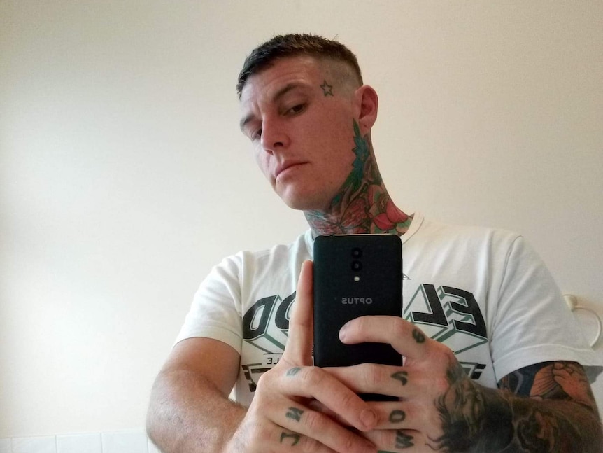 A man with lots of tattoos takes a selfie in the mirror with a mobile phone.