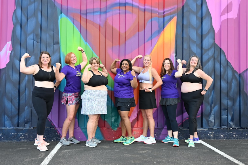 Seven women of all shapes and sizes pose in their sports bras in front of a graffiti wall.