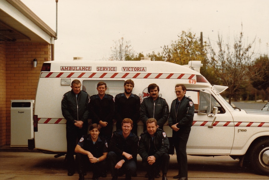 Eight people in paramedics uniforms stand in front of an old-style ambulance.