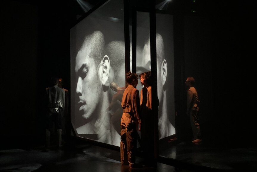 Dancers pause for a moment of stillness on stage, surrounded by large projection screens with hologram-like faces on them.