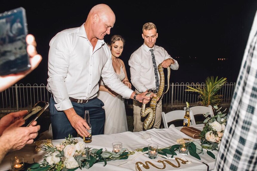 Bridal party guests around table holding snake toward camera.