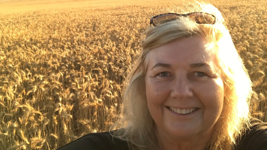 A smiling blonde woman takes a selfie in front of a golden wheat crop.