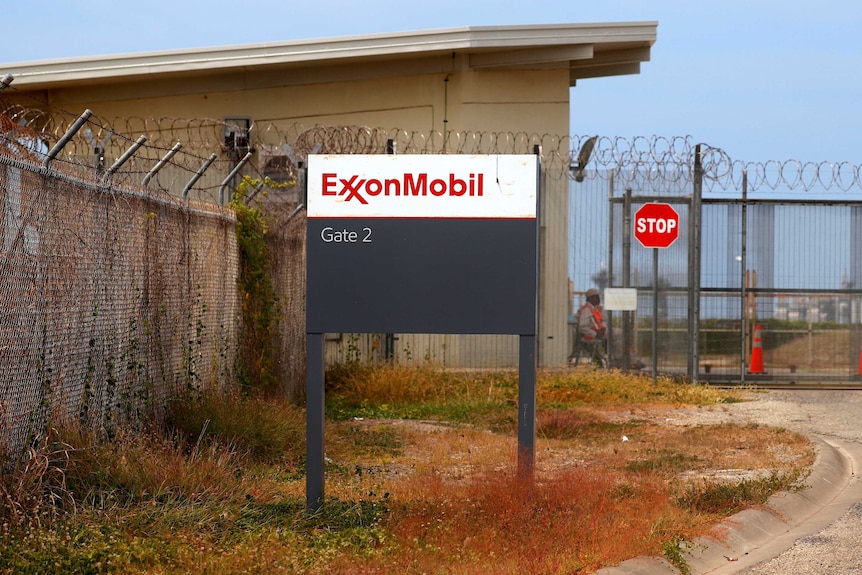 A sign reads Exxon Mobile in front of a stop sign, security guard and building with barbed wire.
