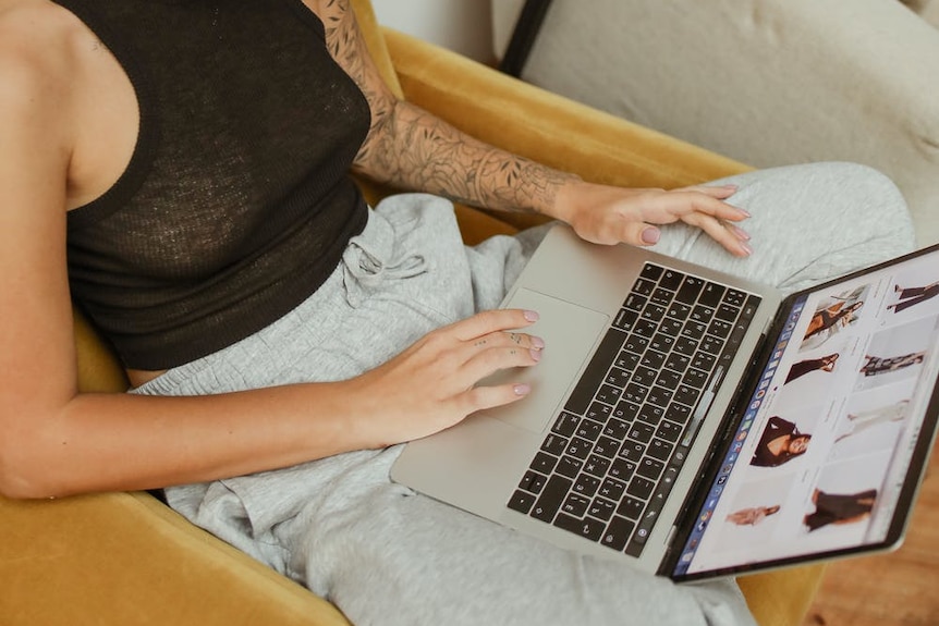 A woman sits on a couch, scrolling through a website while shopping for clothing online. 