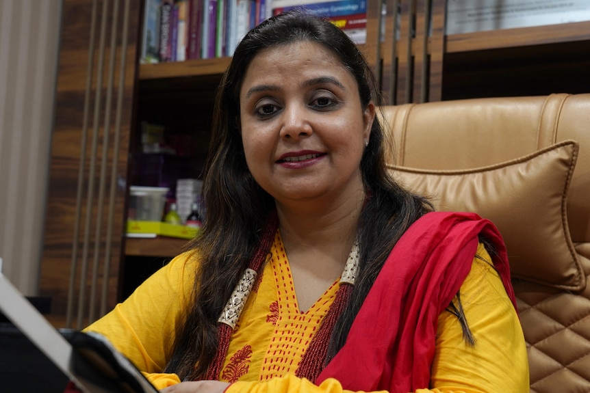 An Indian woman wearing red and yellow clothes sits in front of a bookcase.
