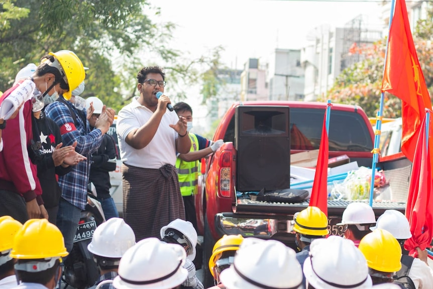 Student organiser Minn Khant Kyaw Linn speaking during a protest with a microphone surrounded by people in hard hats.