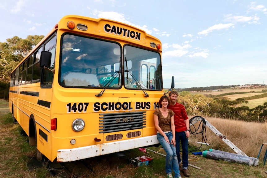 A female and a male stand in front if a large yellow bus which has school bus written on its front