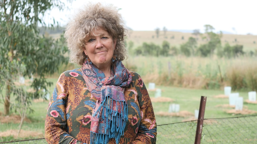 An older woman with curly grey hair stands in front of a paddock.