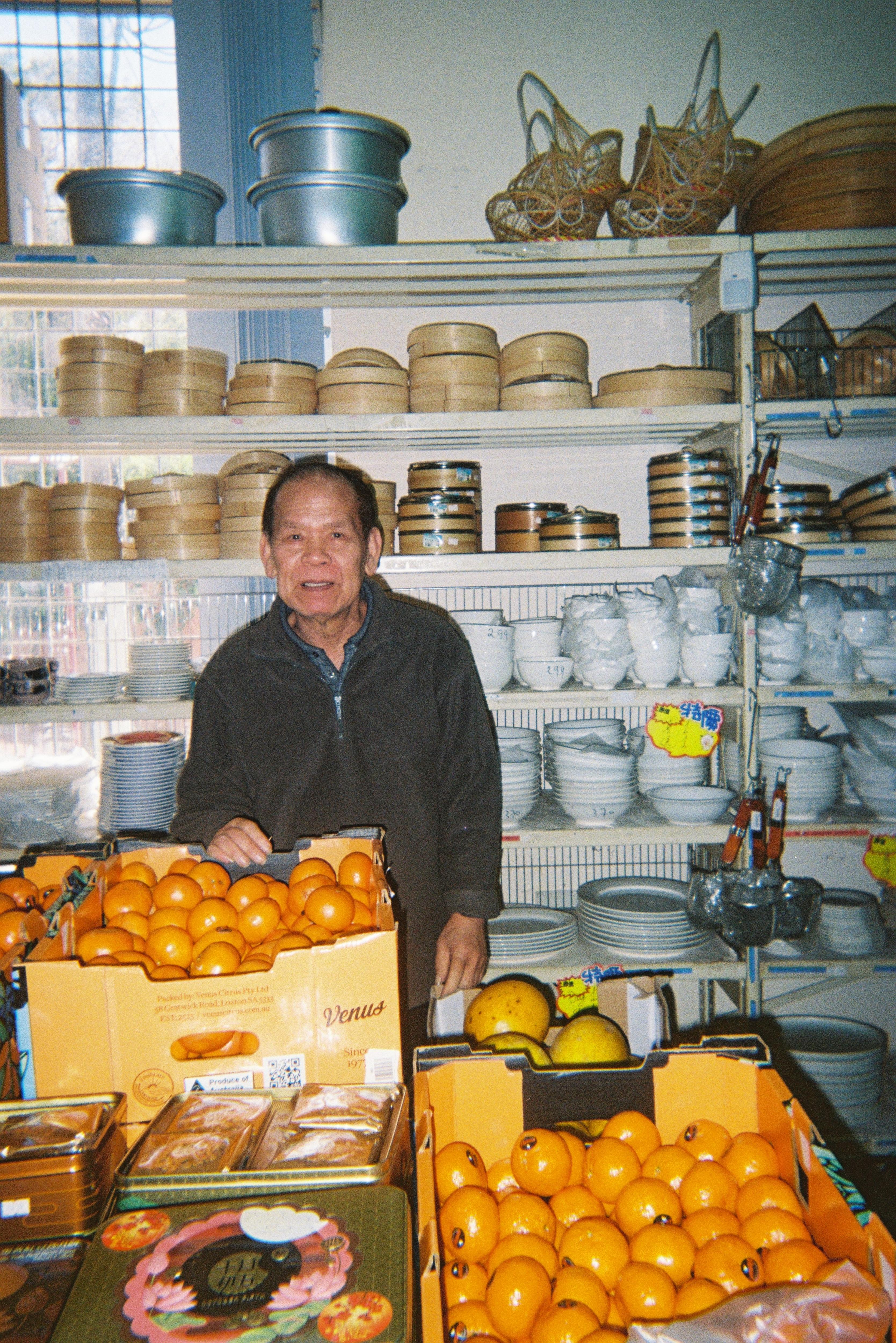 Un hombre asiático se encuentra frente a estantes de vaporeras de bambú y detrás de cajas de cítricos de color naranja.