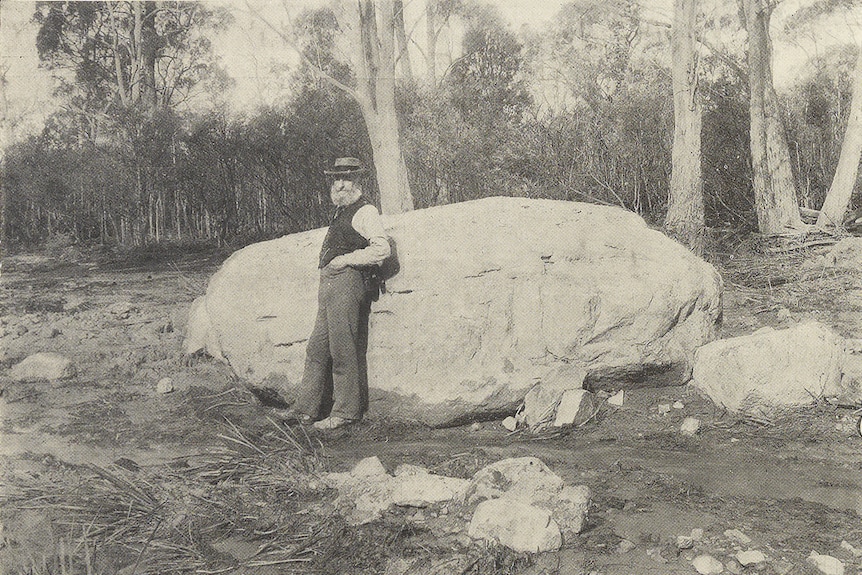 a black and white photo from 1906 of a man standing next to a very large boulder