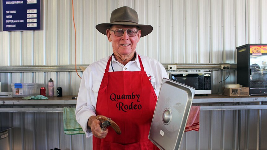 Harold McMillian smiling at the camera, dressed in a red Quamby apron and a cowboy hat, holding a sausage in tongs.