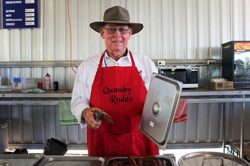 Harold McMillian smiling at the camera, dressed in a red Quamby apron and a cowboy hat, holding a sausage in tongs.