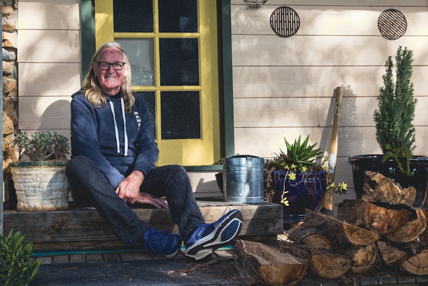 Max sits on the porch of a regional property, next to a pile of chopped wood.