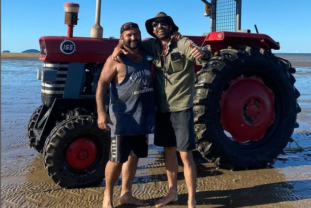 two men stand on the beach next to a tractor