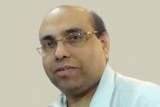 An Indian man with glasses wearing a light white shirt looking at the camera